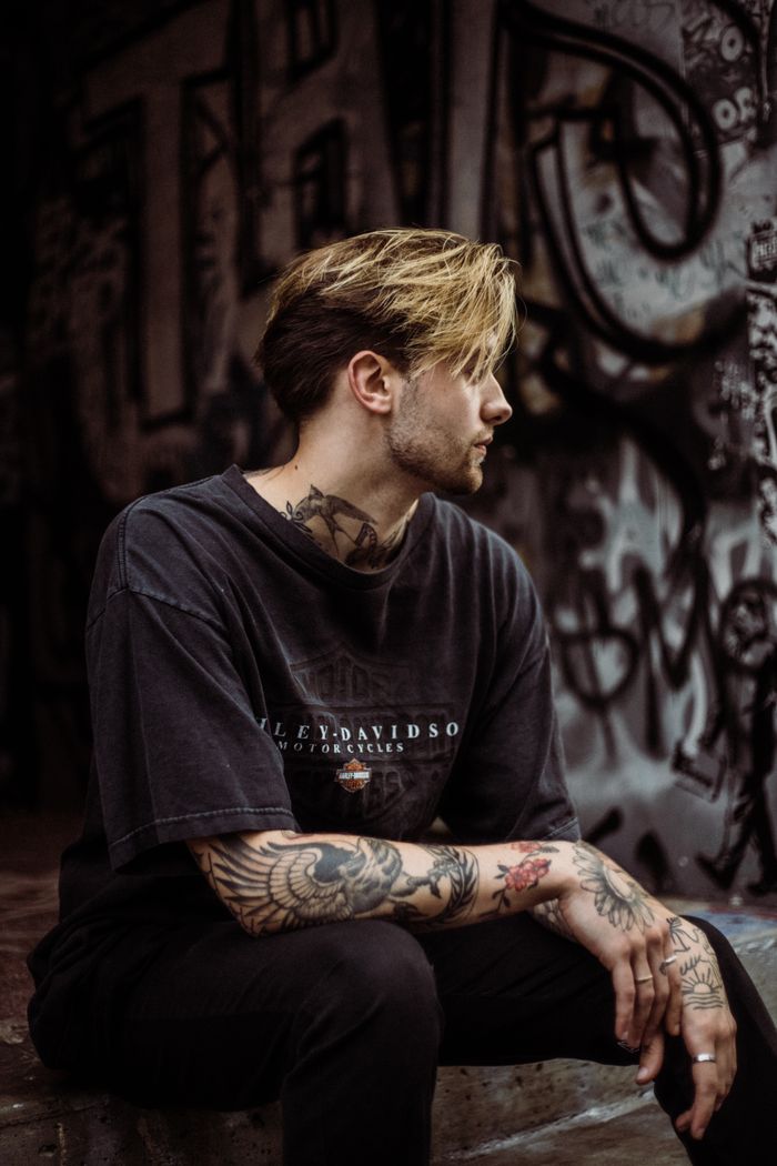 services-03 A tattooed man in a black T-shirt sits in a graffiti-covered urban environment, showcasing street style.